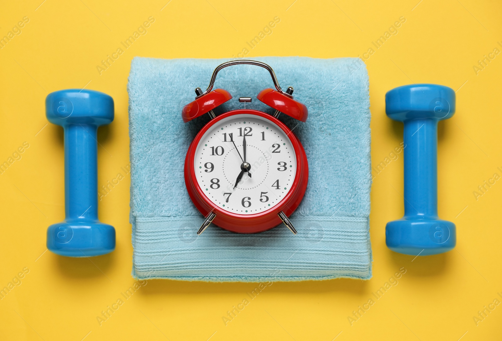 Photo of Alarm clock, towel and dumbbells on yellow background, flat lay. Morning exercise