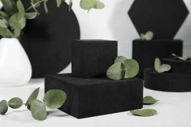 Photo of Black geometric figures and eucalyptus leaves on white marble table, closeup. Stylish presentation for product