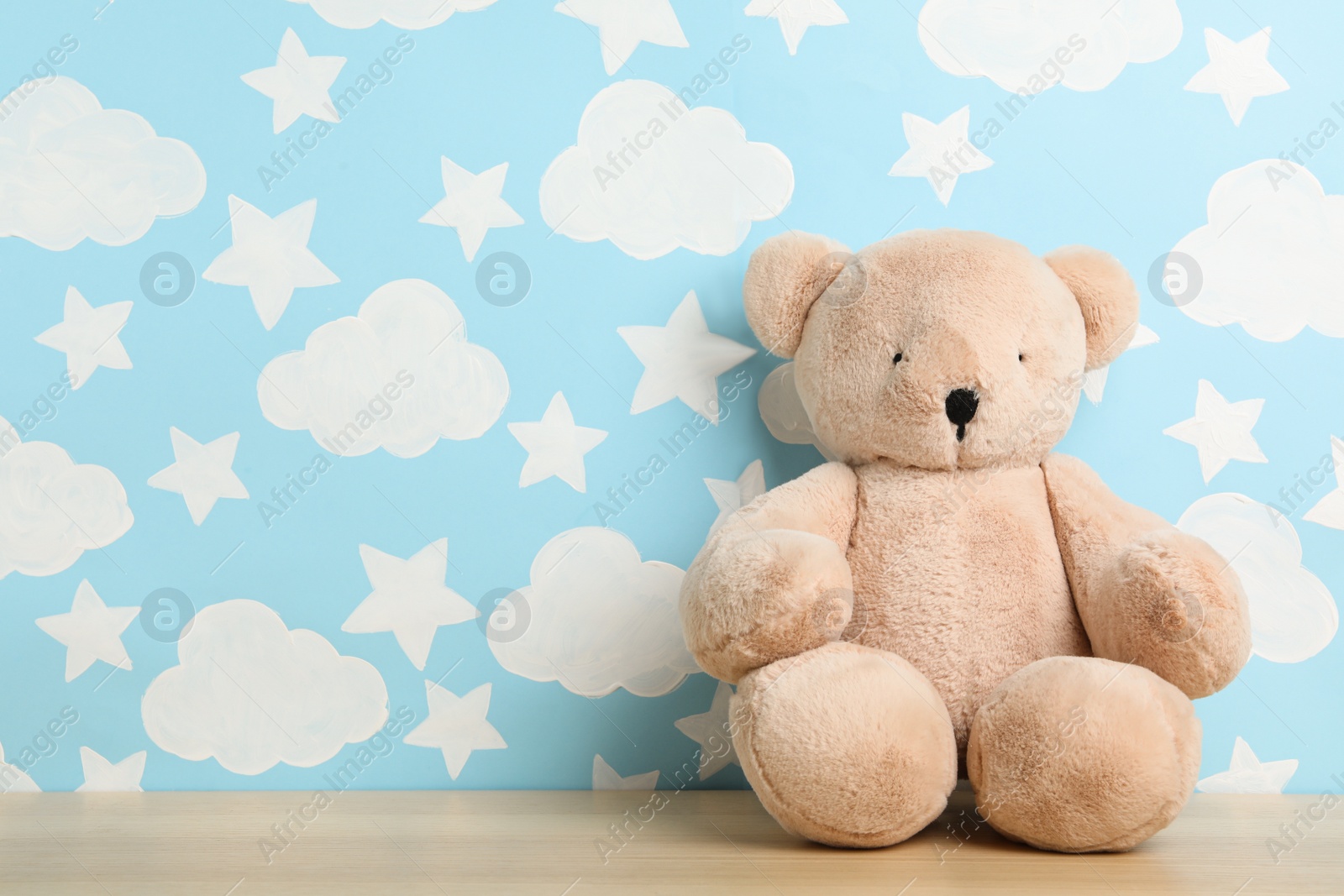 Photo of Teddy bear on wooden table near wall with painted blue sky, space for text. Baby room interior