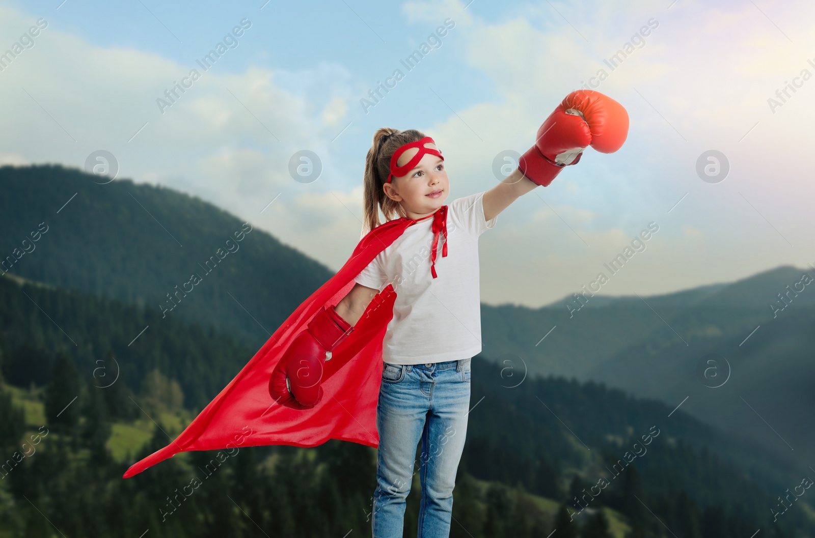 Image of Superhero, motivation and power. Cute girl in cape and boxing gloves on high top in mountains