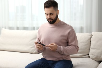 Photo of Diabetes test. Man checking blood sugar level with lancet pen on sofa at home