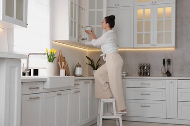 Photo of Woman on ladder putting white jar into cupboard at home