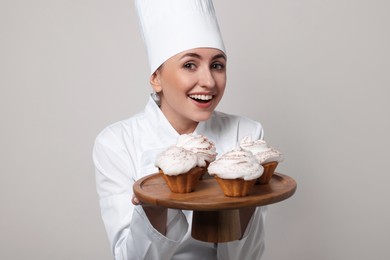 Photo of Happy professional confectioner in uniform holding delicious cupcakes on light grey background