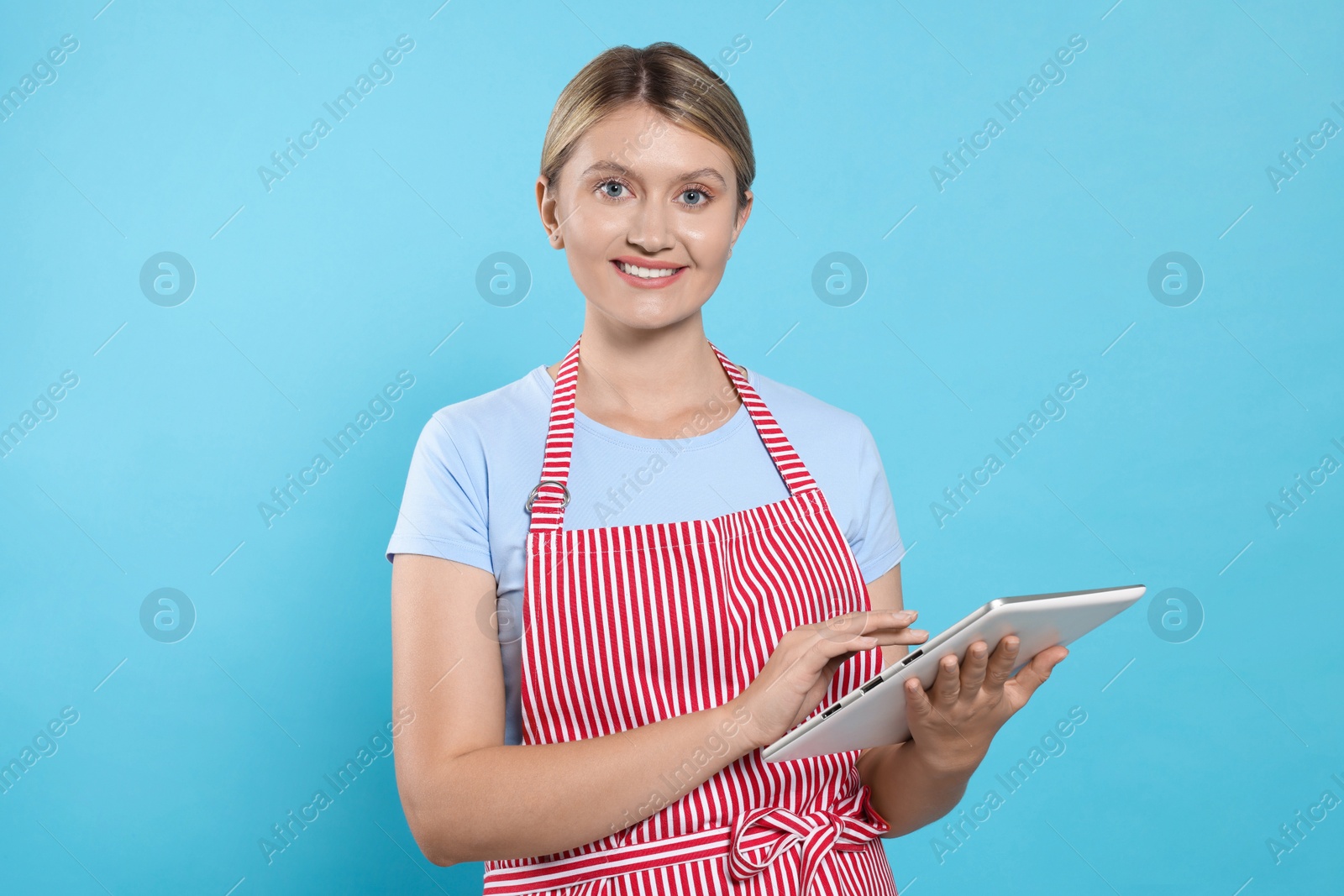 Photo of Beautiful young woman in clean striped apron with tablet on light blue background