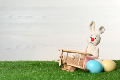 Wooden airplane with cute Easter bunny toy and dyed eggs on green grass, space for text