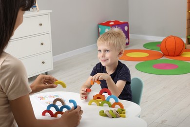 Photo of Motor skills development. Mother helping her son to play with colorful wooden arcs at white table in room