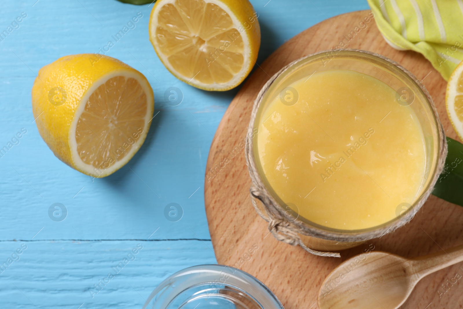 Photo of Delicious lemon curd in glass jar, fresh citrus fruit and spoon on light blue wooden table, top view