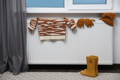 Photo of Heating radiator with knitted hat, sweater, gloves and rubber boots near window indoors