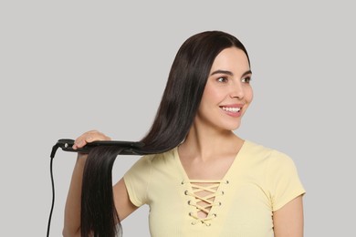 Beautiful happy woman using hair iron on light grey background