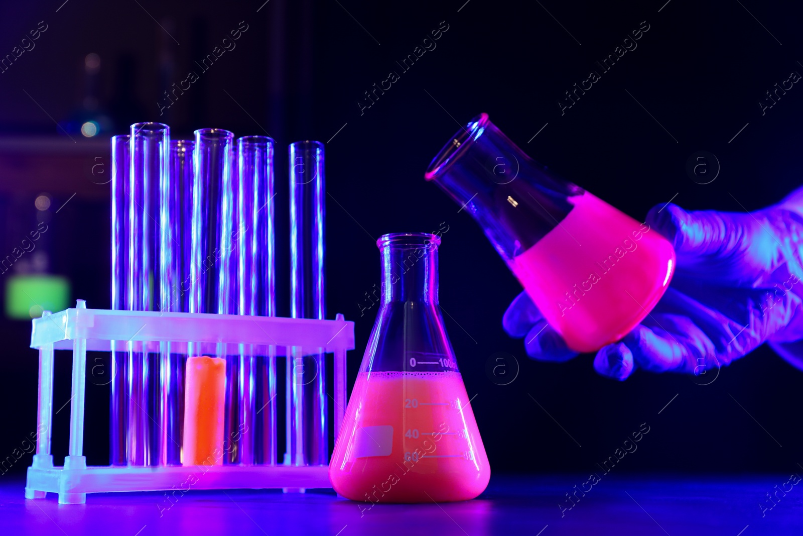 Photo of Scientist working with laboratory glassware of luminous liquids at table against dark background, closeup