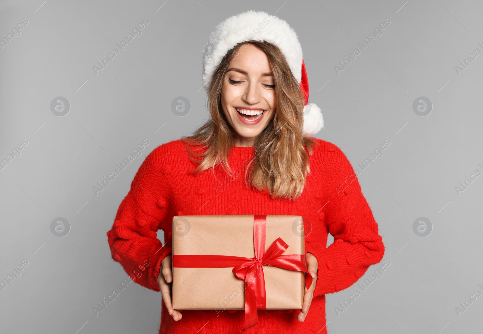 Photo of Happy young woman in Santa hat and sweater with gift box on light grey background. Christmas celebration