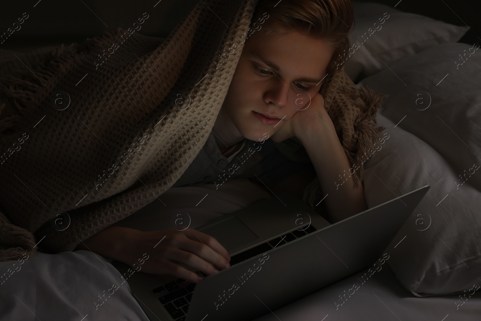 Photo of Teenage boy using laptop under blanket on bed at night. Internet addiction