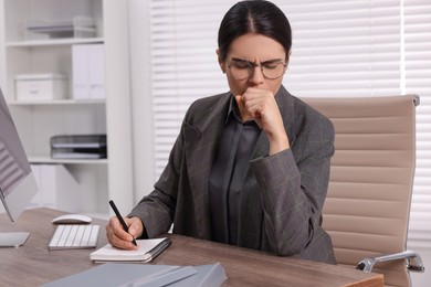 Photo of Woman coughing while taking notes at table in office. Cold symptoms