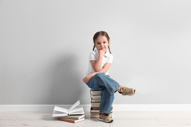 Cute little girl sitting on stack of books near light grey wall