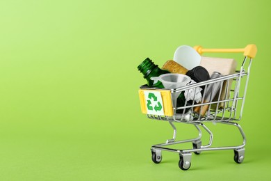 Photo of Shopping cart with recycling symbol full of garbage on light green background, space for text