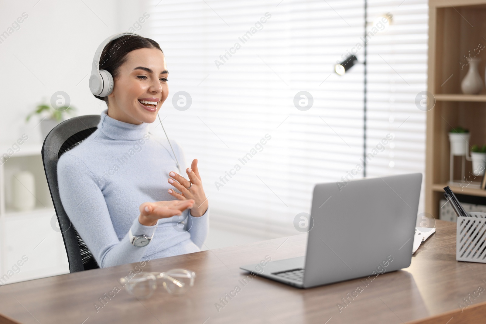 Photo of Young woman in headphones using video chat during webinar at table in office