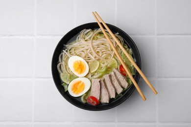 Photo of Bowl of delicious rice noodle soup with meat and egg on white tiled table, top view
