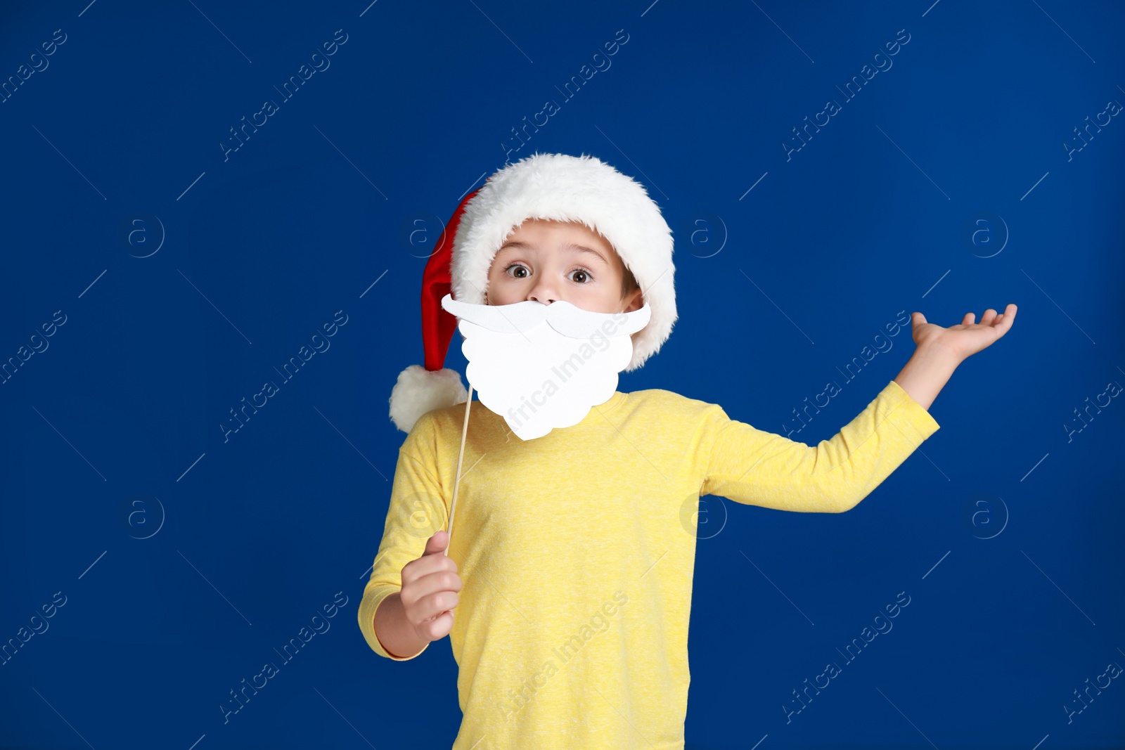 Image of Cute little boy with Santa hat and white beard prop on blue background. Christmas celebration