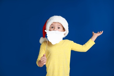 Image of Cute little boy with Santa hat and white beard prop on blue background. Christmas celebration