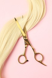 Professional hairdresser scissors with blonde hair strand on pink background, top view