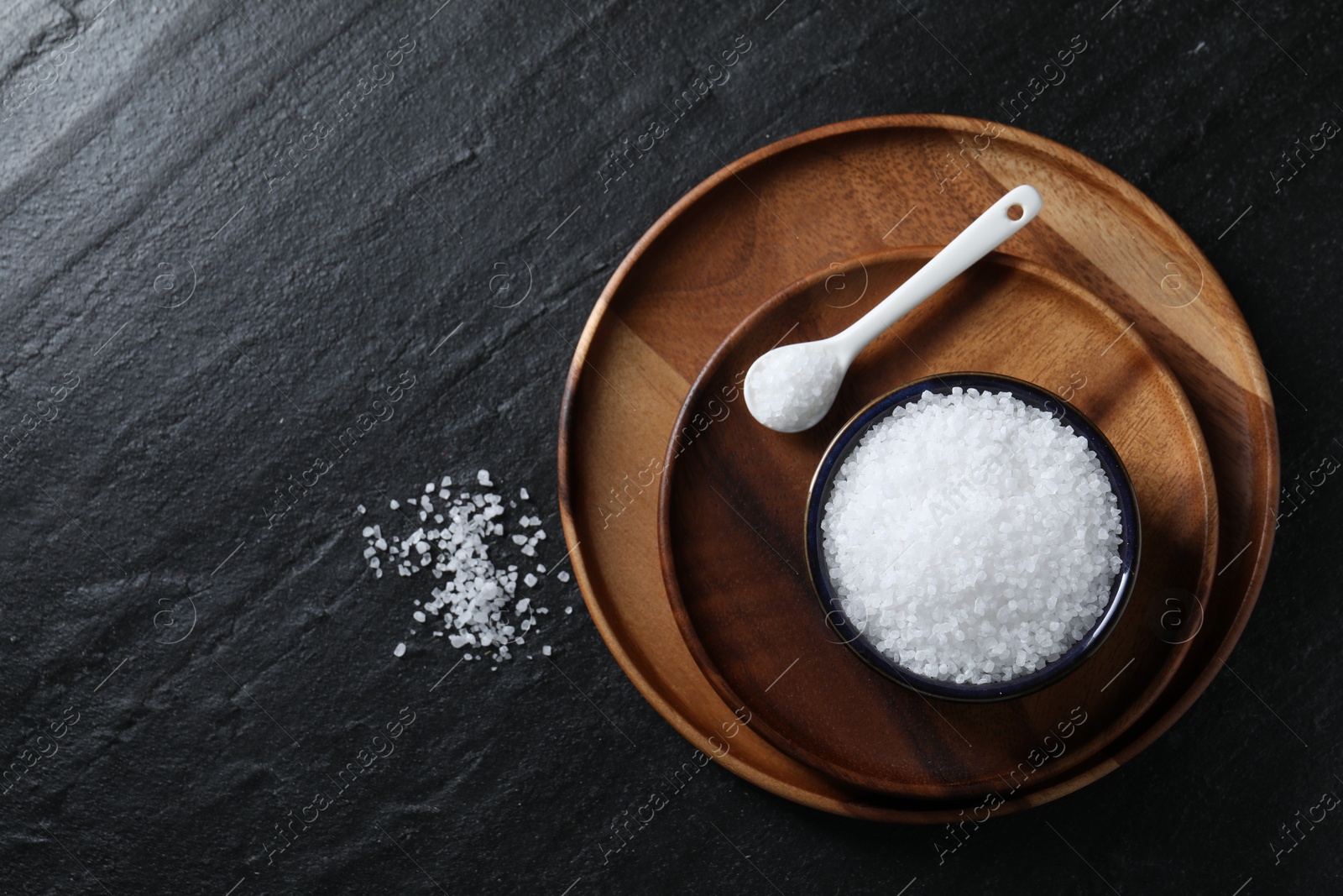 Photo of Organic white salt in bowl and spoon on black table, top view. Space for text