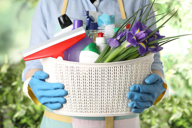 Photo of Woman holding basket with spring flowers and cleaning supplies on green blurred background, closeup