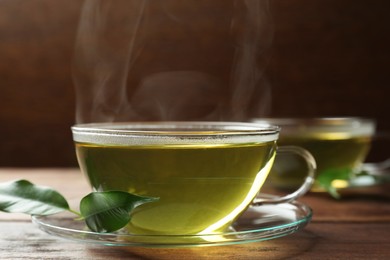 Photo of Fresh green tea in glass cup with saucer and leaves on wooden table, closeup