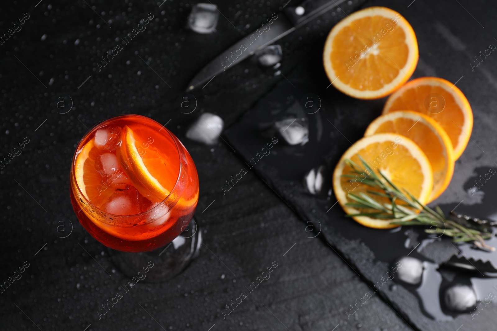 Photo of Glass of tasty Aperol spritz cocktail with orange slices, ice cubes and rosemary on black table, above view