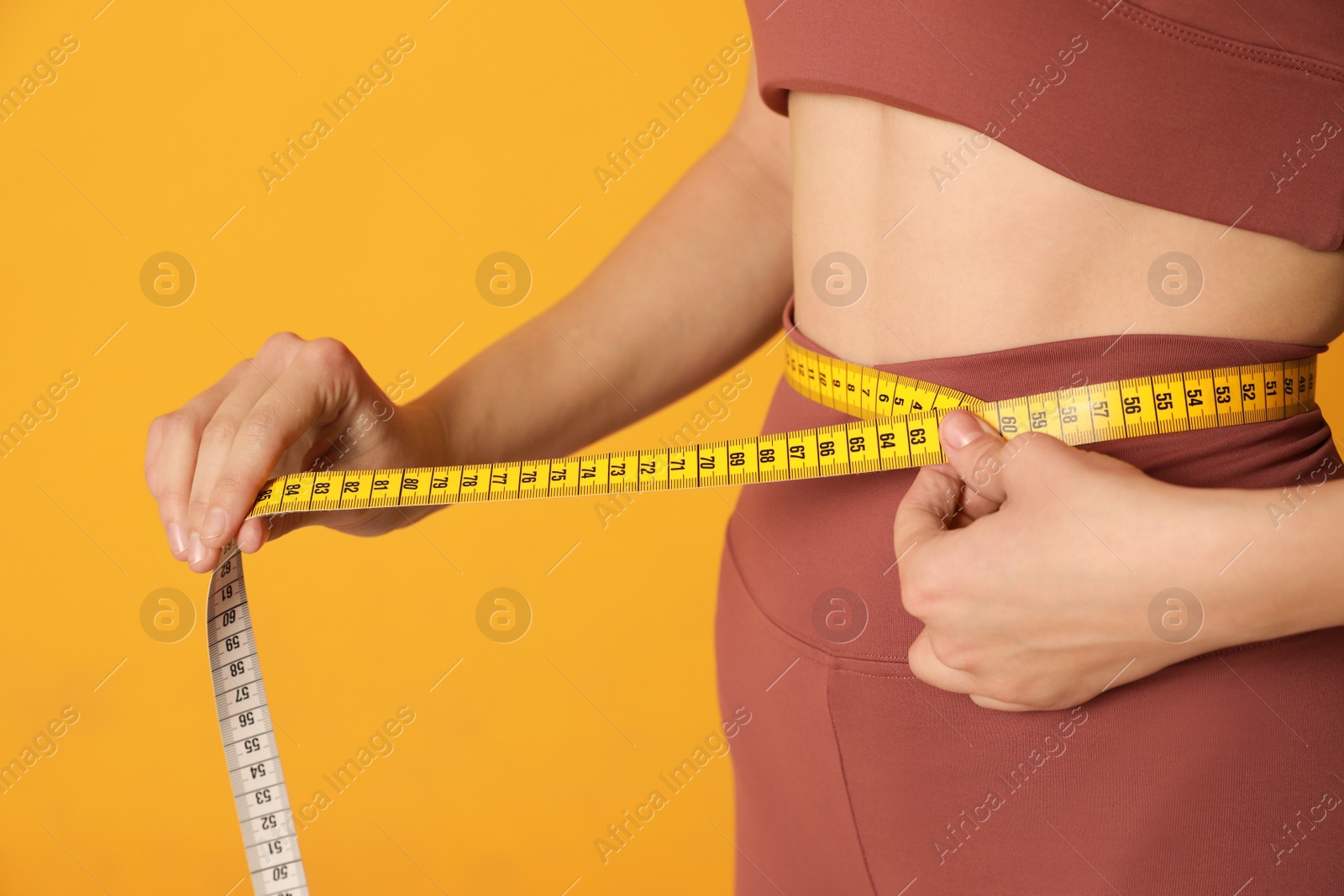 Photo of Woman in sportswear measuring waist with tape on yellow background, closeup