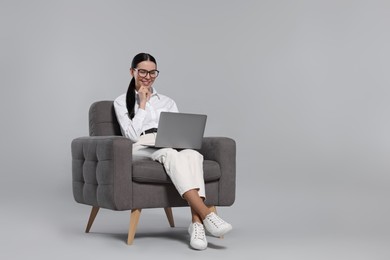 Happy woman with laptop sitting in armchair on light gray background