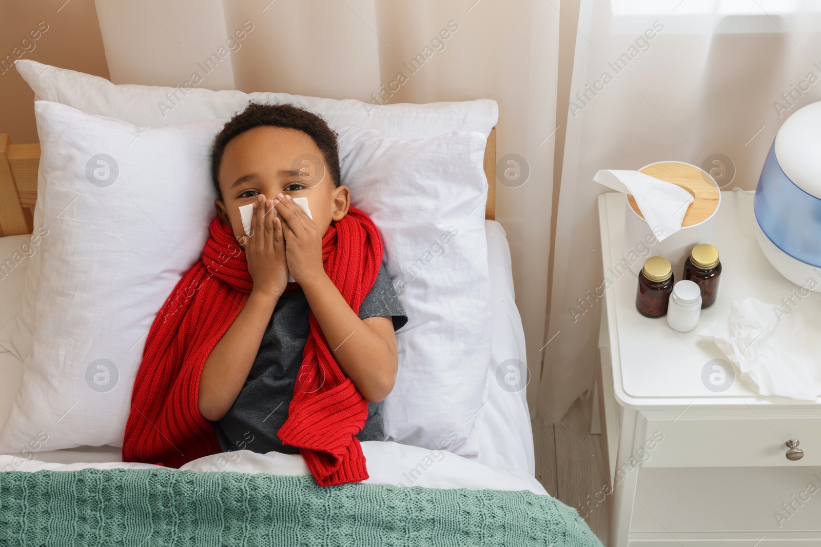 Photo of African-American boy with scarf and tissue blowing nose in bed indoors, above view. Cold symptoms