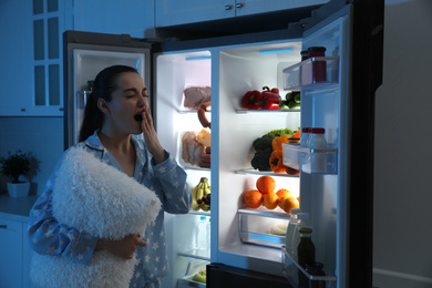 Young woman with pillow near open refrigerator at night