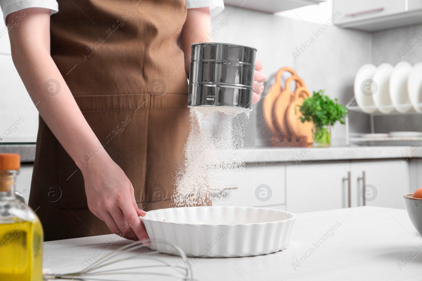 Photo of Woman sieving flour into baking dish at table in kitchen, closeup