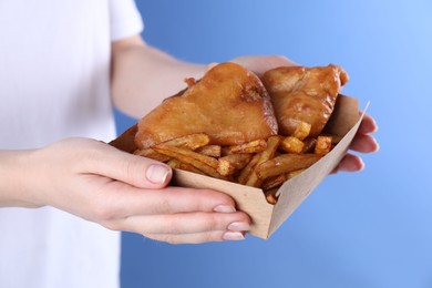 Woman holding fish and chips in paper box on blue background, closeup