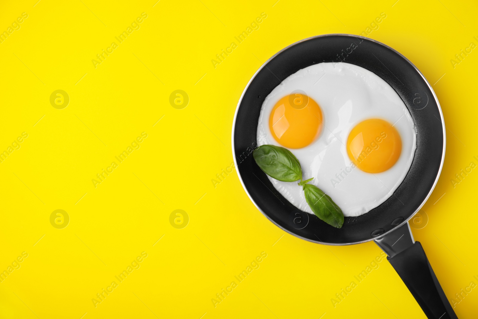 Photo of Tasty fried eggs with basil in pan on yellow background, top view. Space for text
