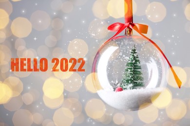 Image of Text Hello 2022 and beautiful transparent Christmas ornament against blurred festive lights, bokeh effect