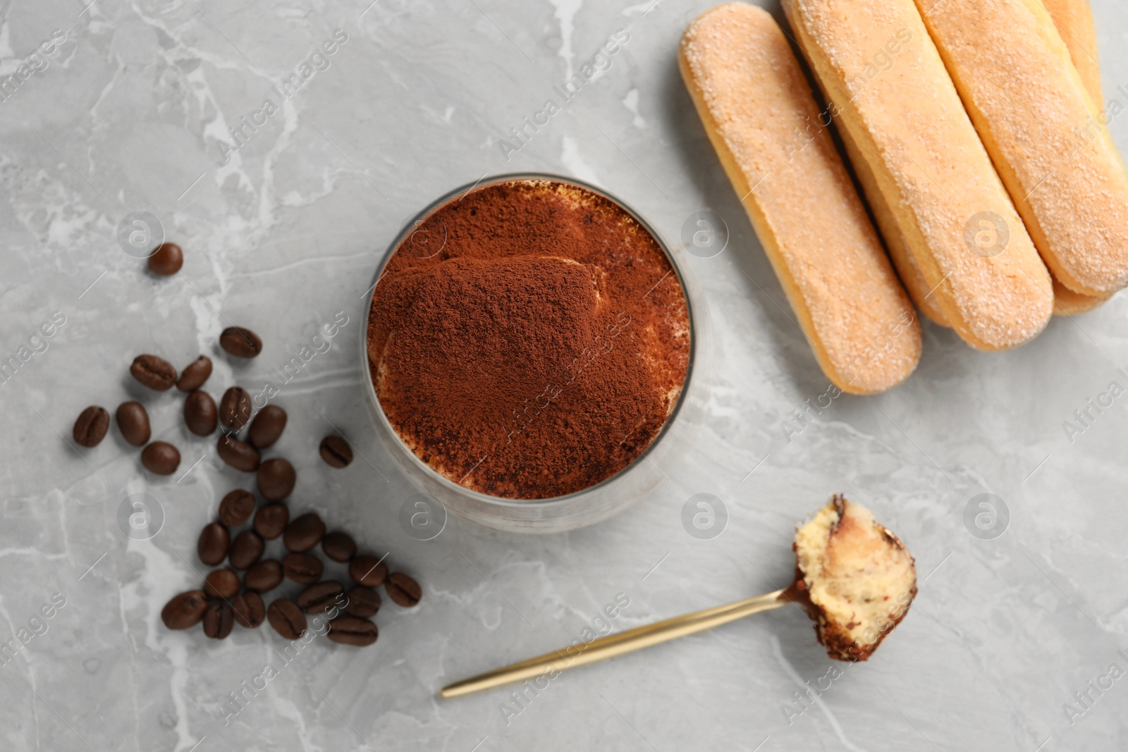 Photo of Tasty tiramisu in glass, coffee beans, spoon and biscuits on light grey table, flat lay