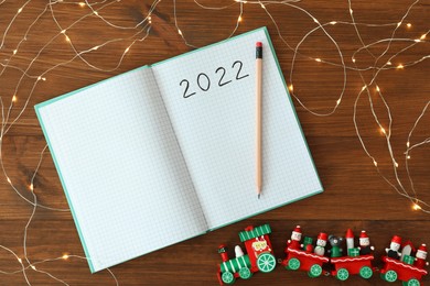 Photo of Open planner, pencil, Christmas lights and toy train on wooden background, flat lay. 2022 New Year aims