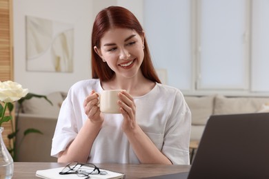 Happy woman with cup of drink using laptop at wooden table in room