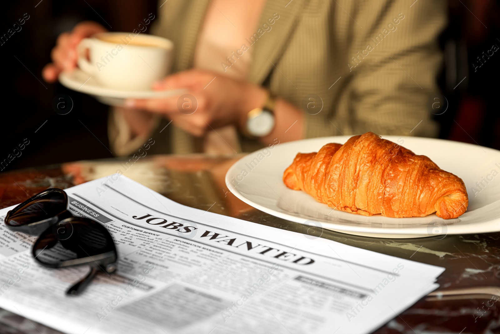 Photo of Woman sitting with cup of hot drink at table, focus on croissant