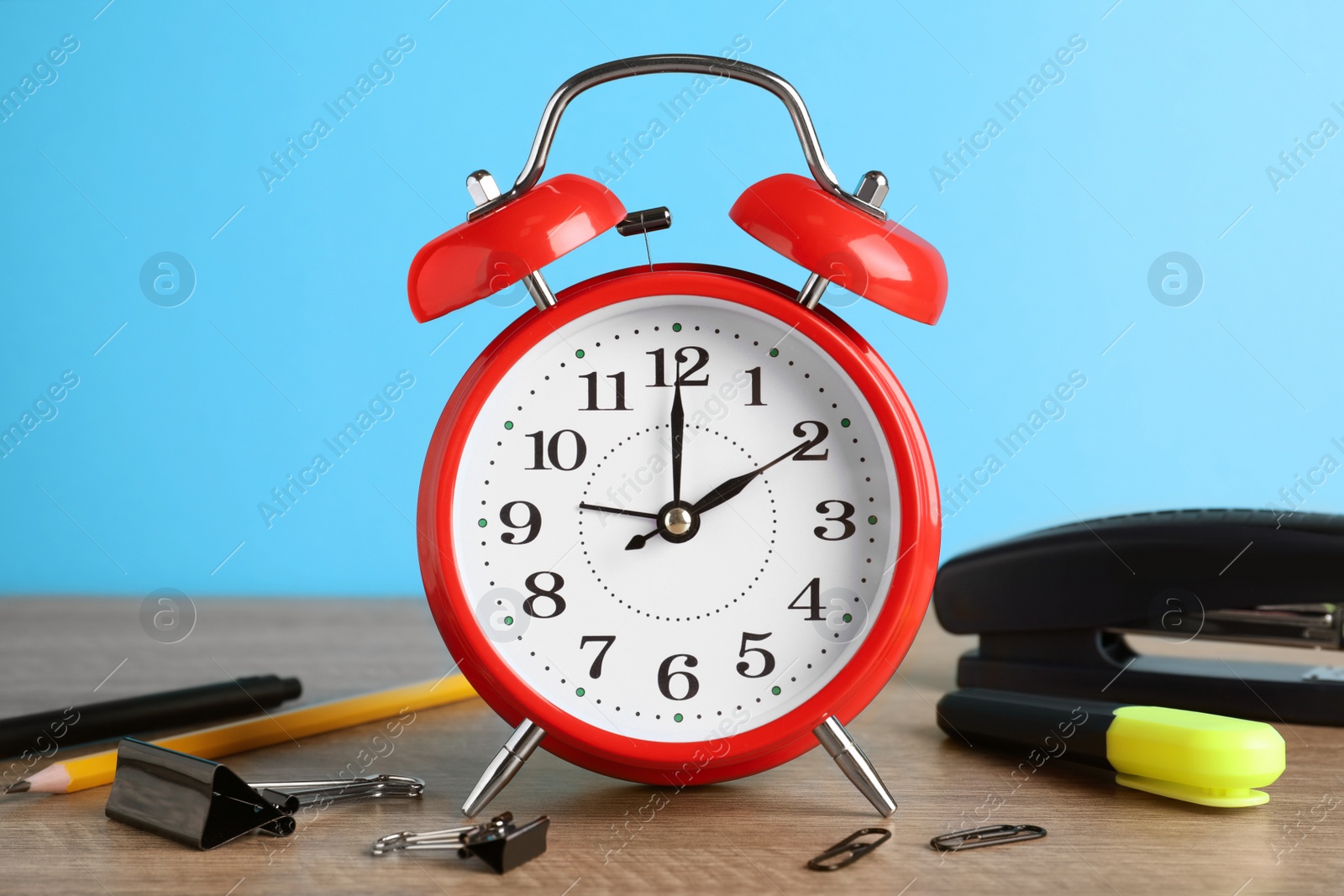 Photo of Red alarm clock and different stationery on wooden table against light blue background. School time