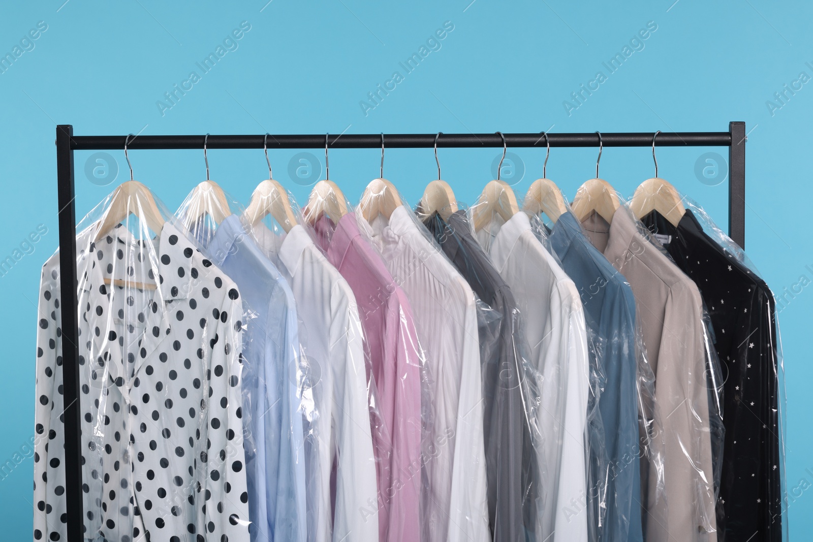 Photo of Dry-cleaning service. Many different clothes in plastic bags hanging on rack against light blue background