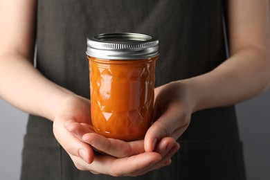 Photo of Woman holding glass jar of delicious persimmon jam on gray background, closeup