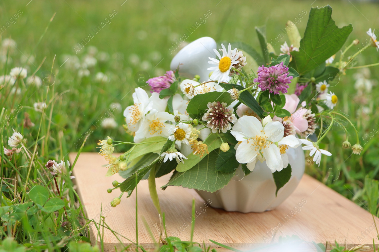 Photo of Ceramic mortar with pestle, different wildflowers and herbs on wooden board in meadow