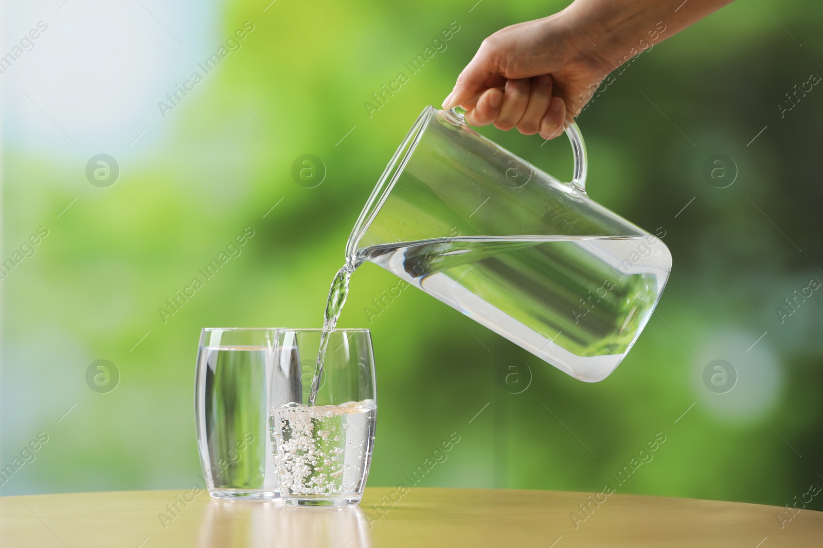 Photo of Woman pouring fresh water from jug into glass at wooden table against blurred green background, closeup