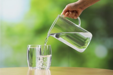 Woman pouring fresh water from jug into glass at wooden table against blurred green background, closeup