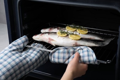 Photo of Woman putting rack with sea bass fish, lemon and thyme into oven, closeup