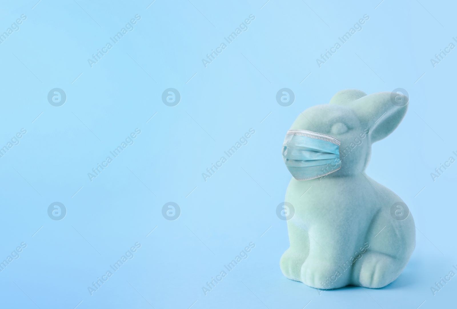 Image of COVID-19 pandemic. Easter bunny toy in protective mask on light blue background, space for text 