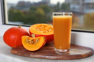 Photo of Tasty pumpkin juice in glass, whole and cut pumpkins on windowsill indoors