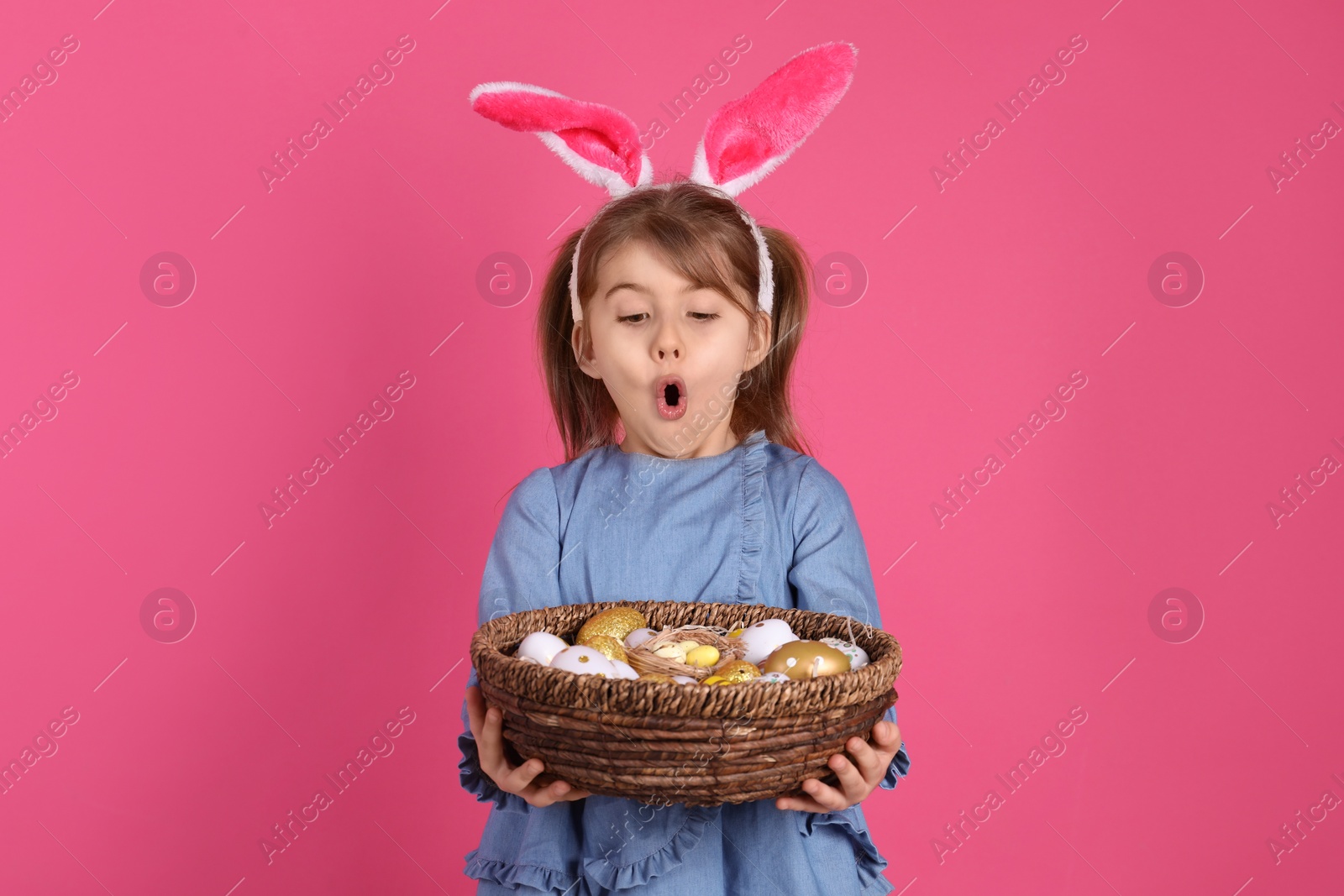 Photo of Surprised little girl with bunny ears holding wicker basket full of Easter eggs on pink background